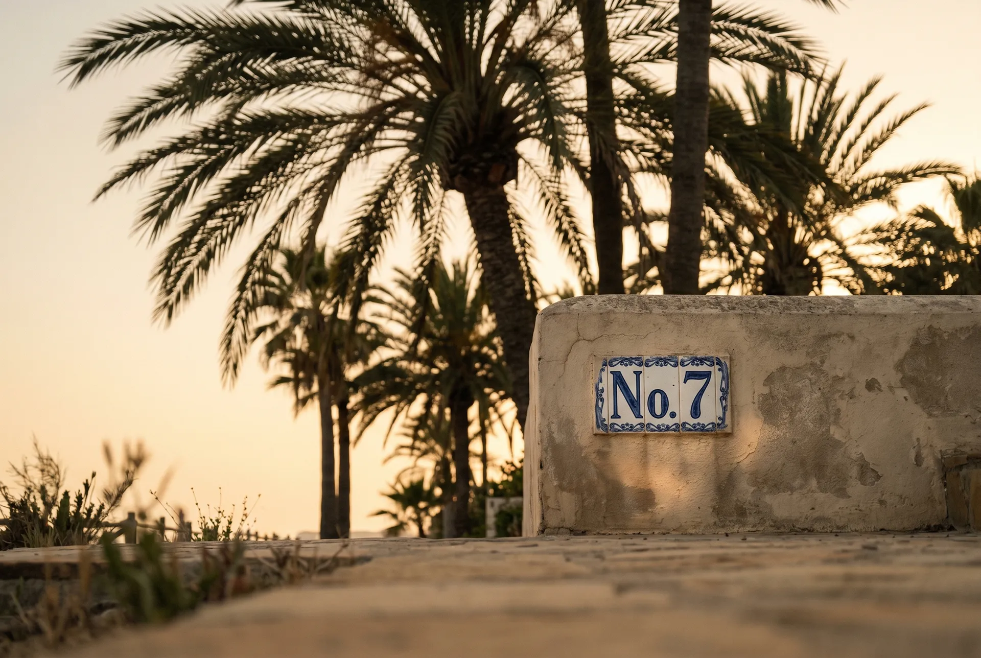 Palm silhouettes at sunset on the Marbella Golden Mile with a fragment of an older boutique-style villa wall and hand-painted ceramic numbering reading 