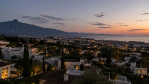 Elevated panoramic view of central Marbella and its bay at blue hour with warm city lights starting to glow and La Concha mountain in soft silhouette, framing the 2026 Marbella property market for international buyers