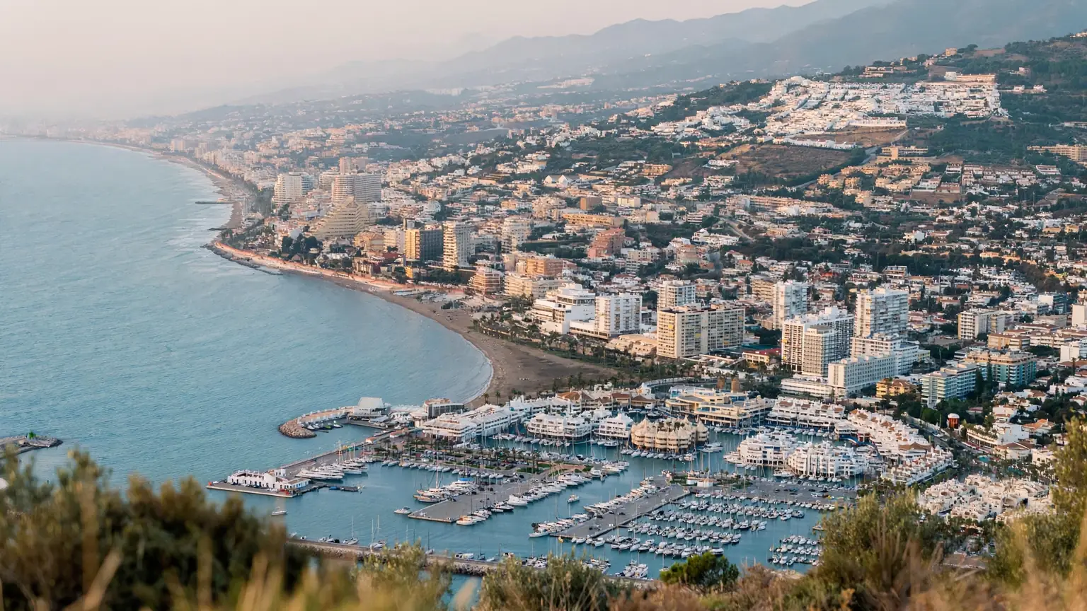 Elevated view over Benalmadena with the marina and dense coastal strip below and the residential hillside climbing toward the mountains behind