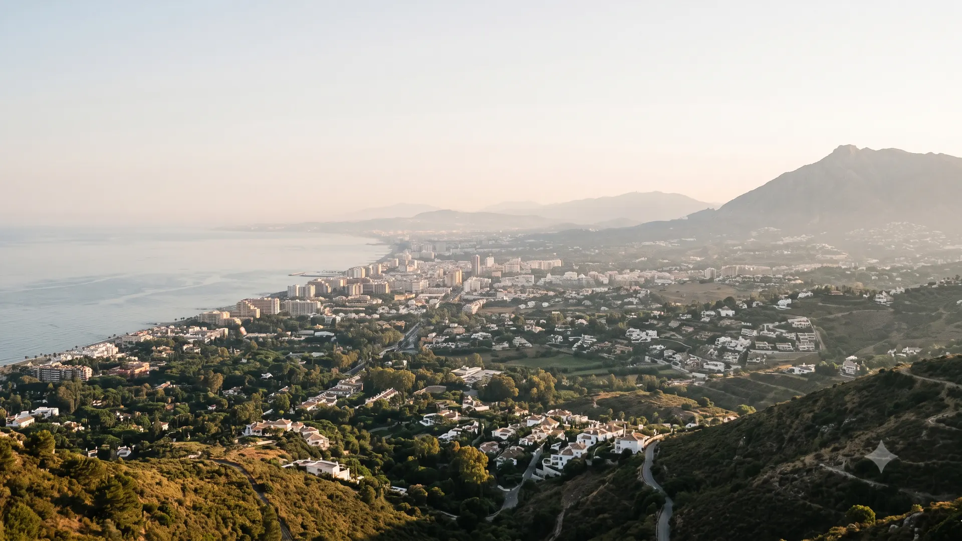 Elevated dusk view along the Marbella coastline, showing the dense town centre, the greener residential hillsides around it and the mountains rising to the east