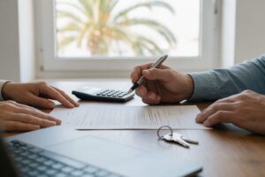 Hands signing a mortgage document next to a calculator, laptop and house keys in a bright Spanish office
